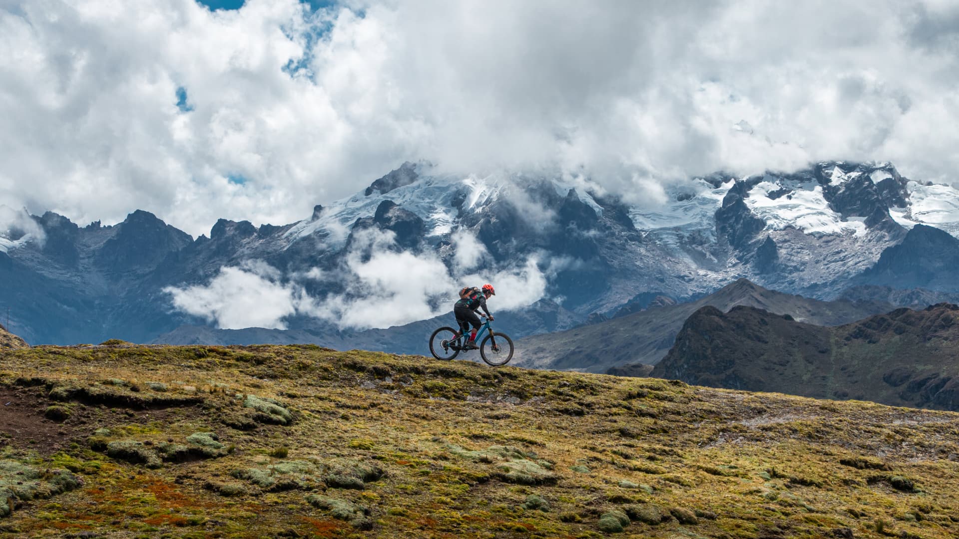Ruta Épica de Ciclismo frente al Nevado Veronica, Cusco Ciclista de montaña recorriendo un sendero en lo alto de los Andes, con el imponente glaciar del Apu Veronica cubierto de nubes al fondo.