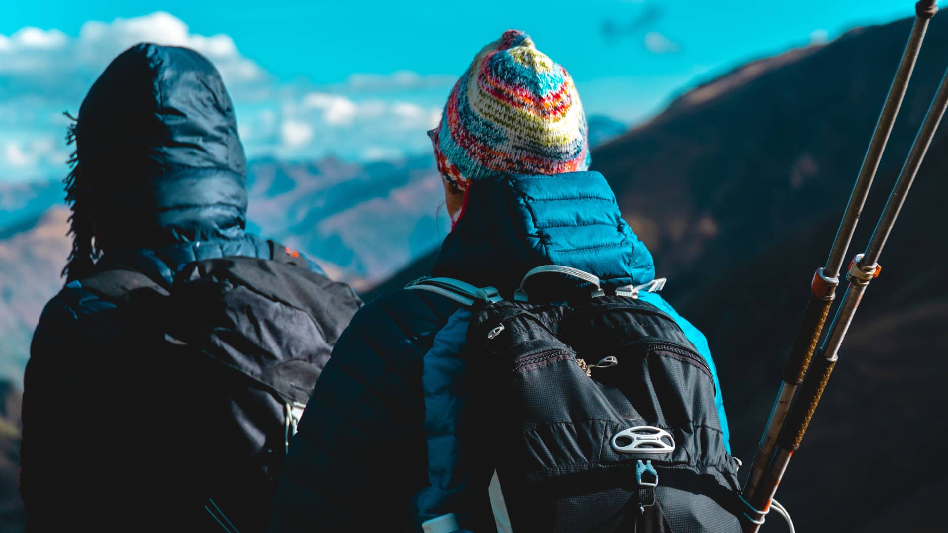 Mirador en los Andes: La Recompensa de tu Aventura Lares Trek Dos excursionistas de espaldas, equipados con mochilas y gorros, hacen una pausa para admirar el vasto paisaje de montañas en lares trek en Perú.