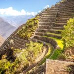 Andenes incas de piedra descienden por una montaña verde y soleada en el sitio arqueológico de Choquequirao, con los Andes de fondo.