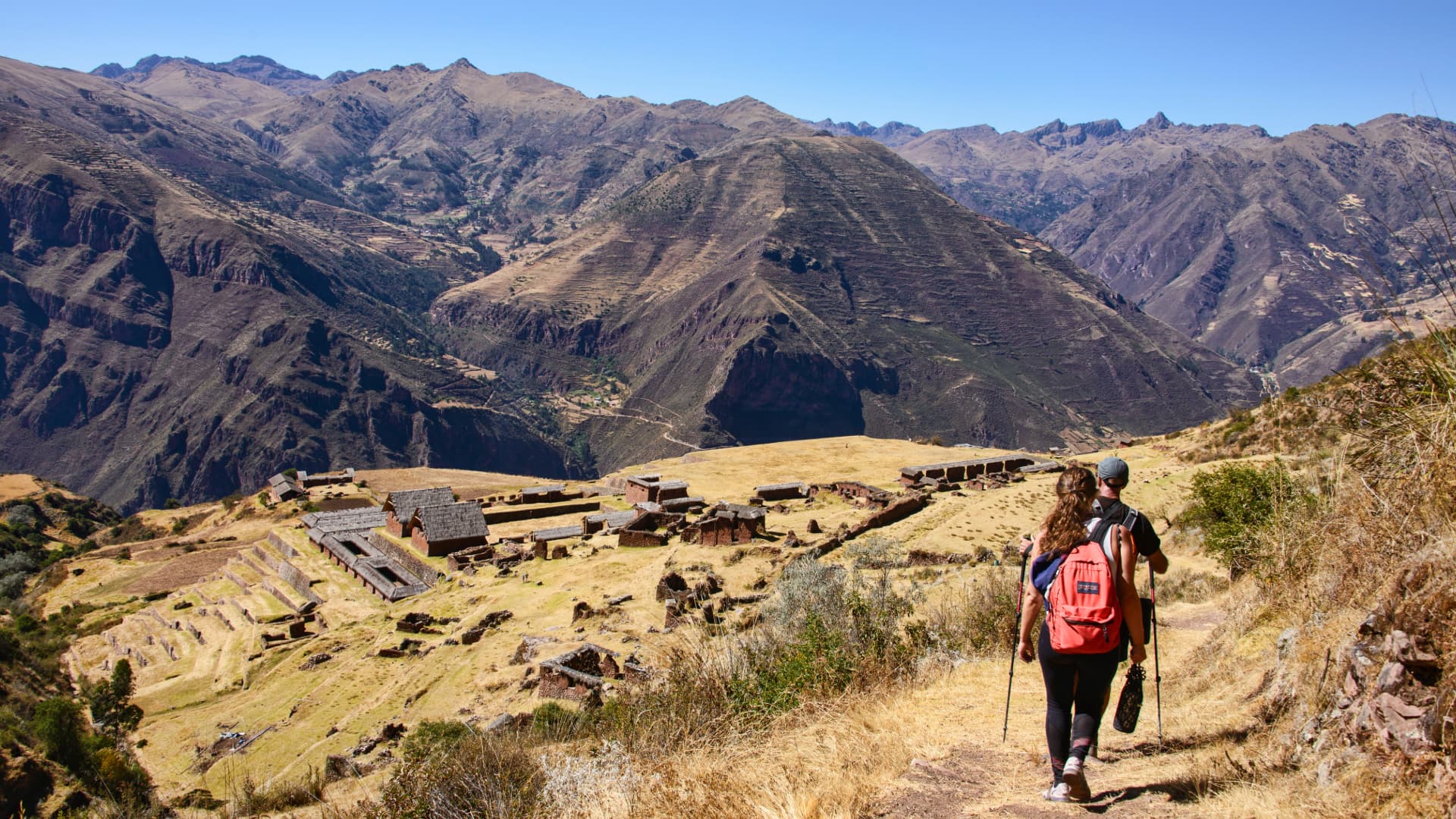 Trek a Huchuy Qosqo: Aventura en el Valle Sagrado, Cusco Dos excursionistas caminando por un sendero andino con vistas a las antiguas ruinas incas de Huchuy Qosqo en el Valle Sagrado.