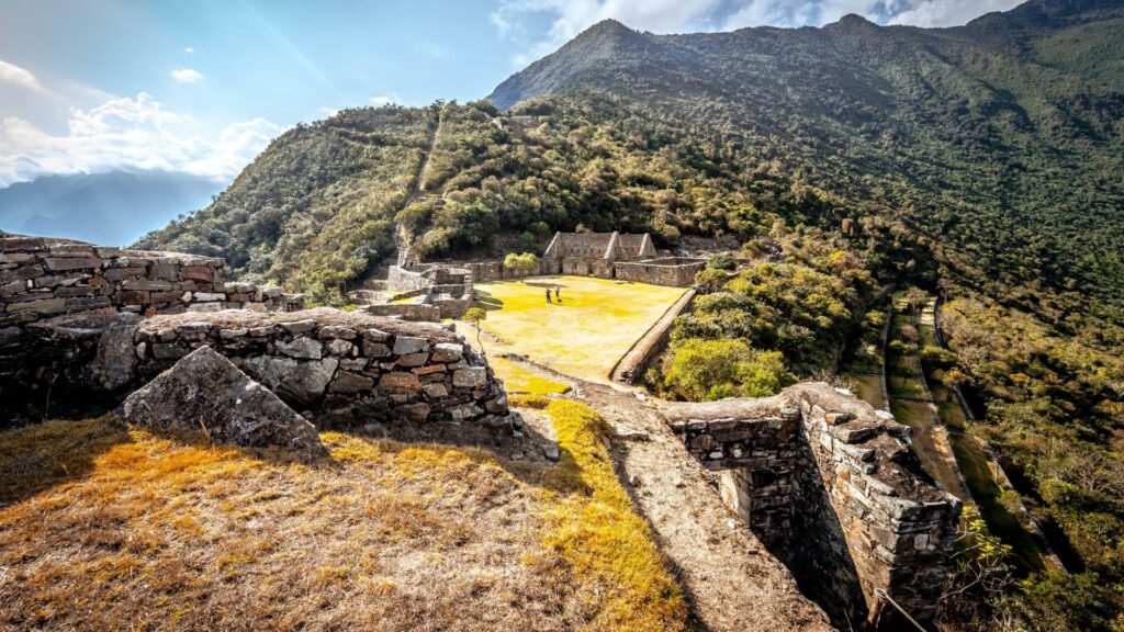 Vista desde las ruinas superiores hacia la plaza principal de Choquequirao, con dos excursionistas en el centro y los Apus andinos de fondo.
